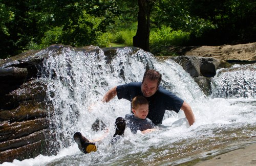 A man and a boy joyfully splash in a waterfall, surrounded by lush greenery on a sunny day.