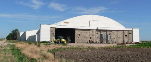 A large, white agricultural building with a curved roof, surrounded by open land and a green tractor in front.