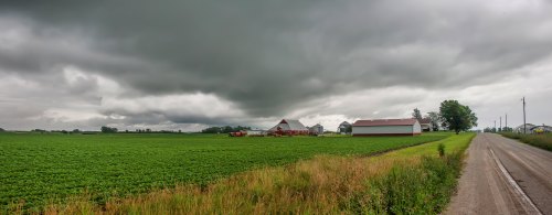 A panoramic view of a farm with green fields, barns, and a cloudy sky, alongside a gravel road.