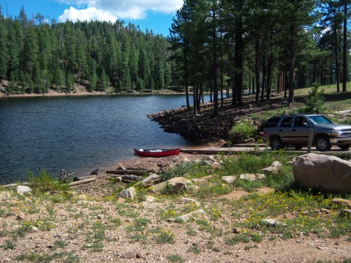 A serene lakeside scene with a red kayak on the shore, surrounded by trees and a parked SUV nearby.