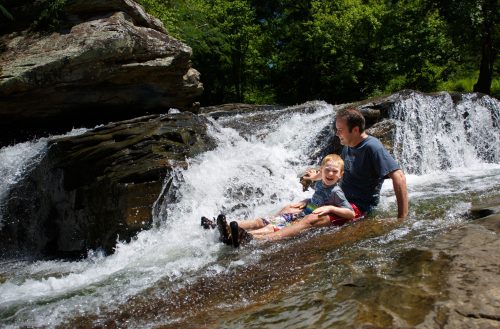A man and a boy sit on a rock by a waterfall, enjoying the splashing water on a sunny day.