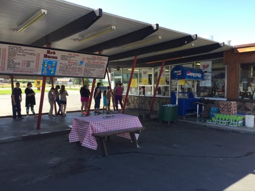 A busy outdoor ice cream stand with a menu board, people in line, and picnic tables under a shaded roof.