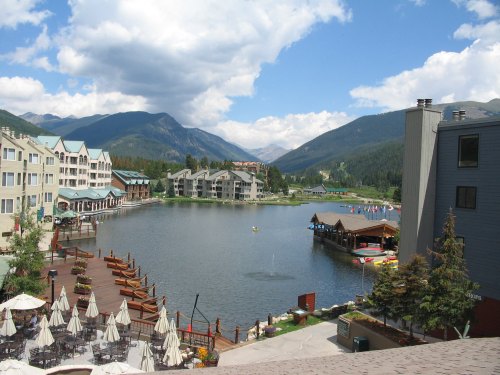 Scenic view of a lake surrounded by mountains, with buildings and boats along the shore under a partly cloudy sky.