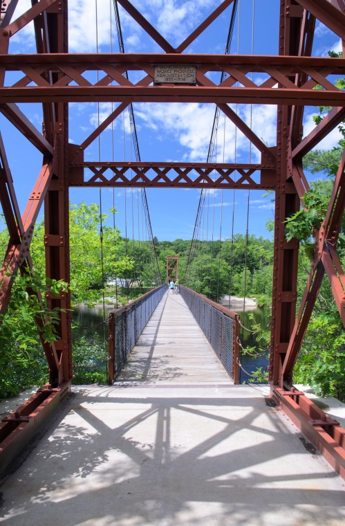 A red suspension bridge spans a river, surrounded by lush greenery and a clear blue sky.