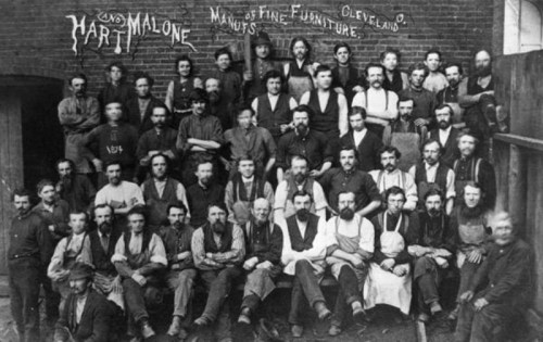 A historic black-and-white photo of a large group of men, likely workers, posing in front of a furniture factory.