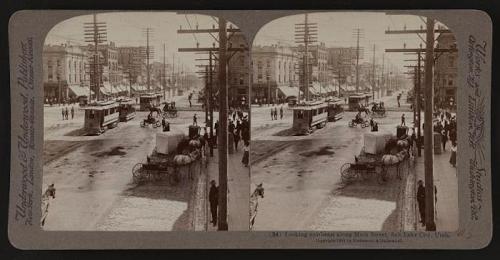 Historic street scene showing horse-drawn carriages and early electric streetcars on a busy city street.