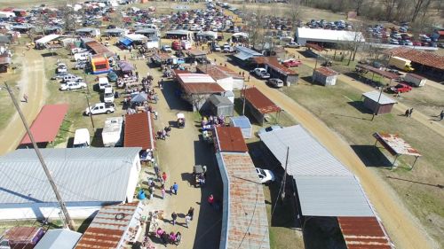 Aerial view of a bustling outdoor market with vendors, parked cars, and people walking along dirt paths.