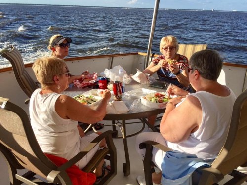 Four women enjoy a meal together on a boat, with water and a clear sky in the background.