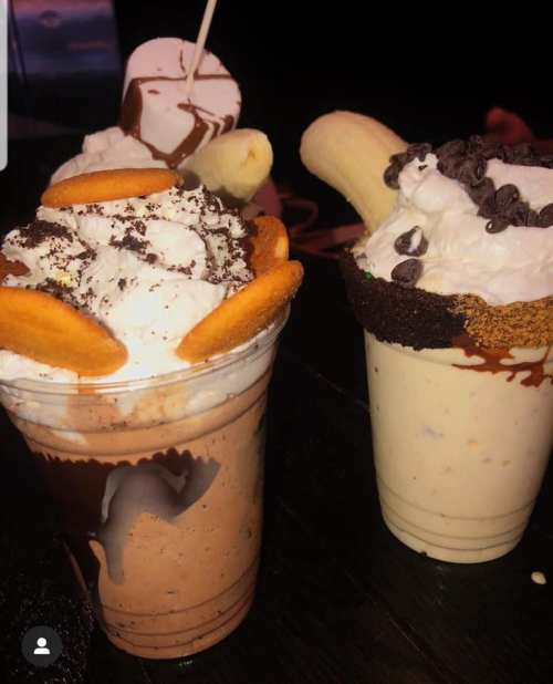 Two decadent milkshakes topped with whipped cream, cookies, and banana slices, served in clear cups.