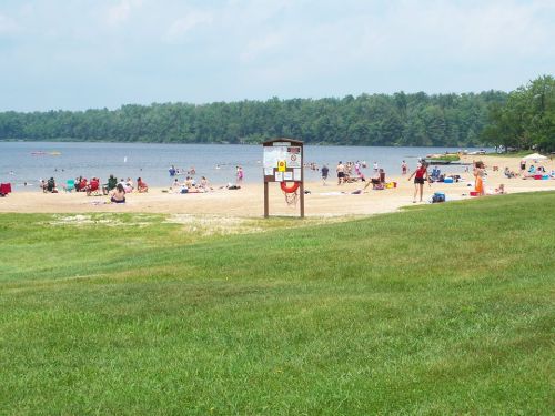 A sandy beach with people sunbathing and swimming, surrounded by green trees and a calm lake. Lifeguard station visible.