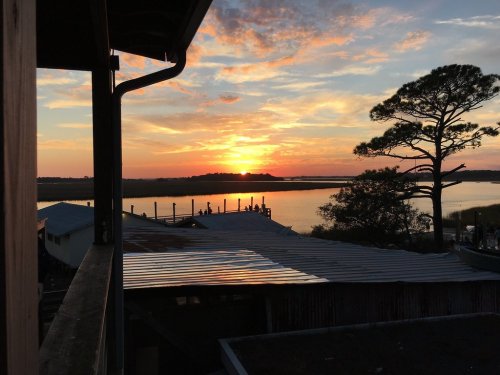 Sunset over a calm waterway, with silhouettes of trees and buildings in the foreground, creating a serene atmosphere.