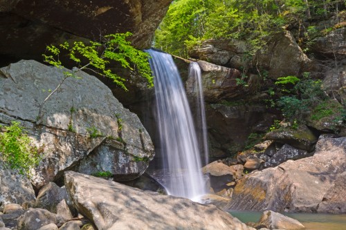 A serene waterfall cascades over rocks, surrounded by lush greenery and towering cliffs in a tranquil natural setting.