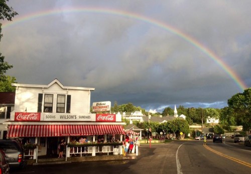 A charming restaurant with a striped awning under a vibrant rainbow and cloudy sky, surrounded by greenery.