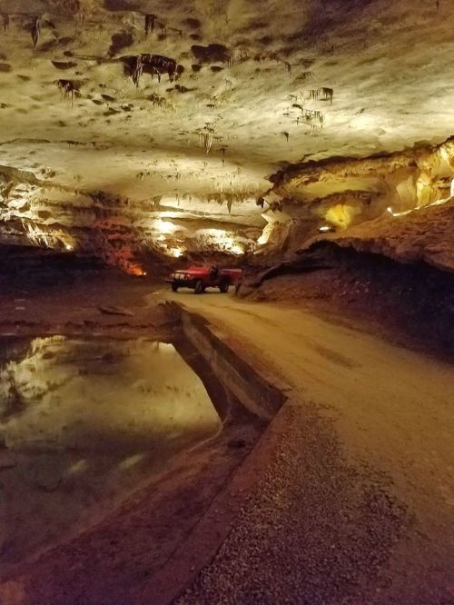 A dimly lit cave interior with rock formations, a still water pool, and a red vehicle parked along a gravel path.