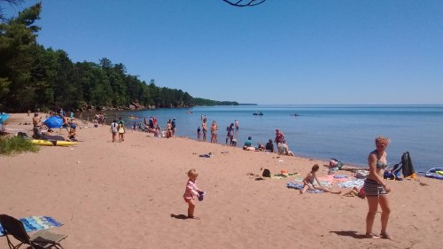 A sunny beach scene with people enjoying the water, children playing, and trees lining the shore.