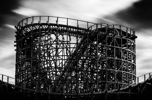 A black and white image of a wooden roller coaster structure against a cloudy sky.