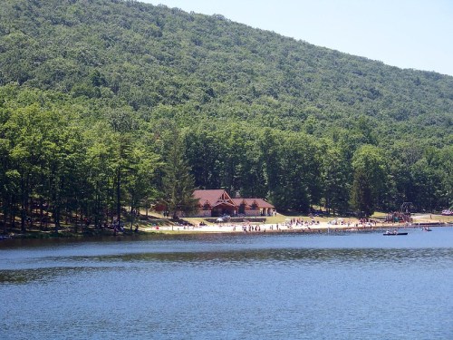 A serene lake surrounded by green trees, with a sandy beach and a lodge in the background, filled with people enjoying the day.