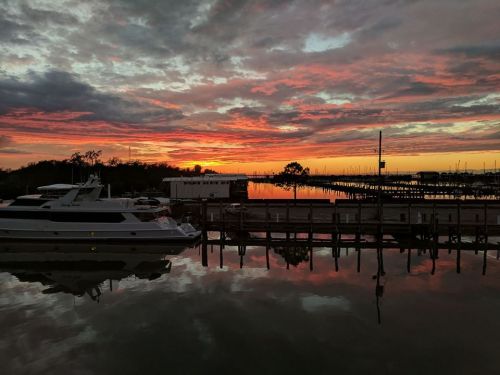 A vibrant sunset reflects on calm waters, with boats docked and a colorful sky filled with clouds.