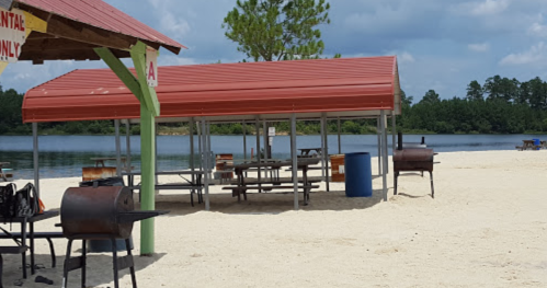 A sandy beach area with a red-roofed shelter, picnic tables, and grills near a calm lake under a partly cloudy sky.