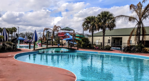 A sunny pool area with water slides, palm trees, and lounge chairs under blue skies and scattered clouds.