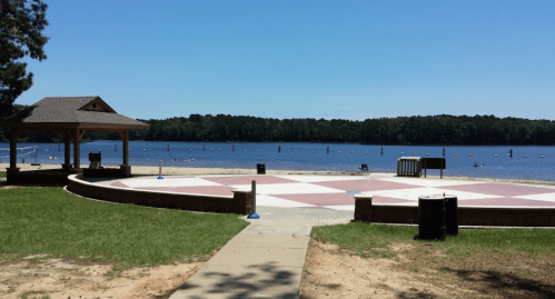 A sunny lakeside view featuring a gazebo, a paved area, and people enjoying the water in the background.