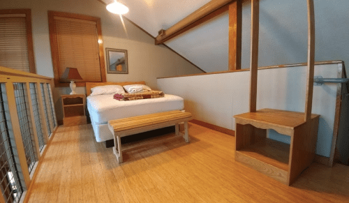 Cozy loft bedroom with a bed, wooden bench, and a lamp, featuring warm wood accents and natural light.