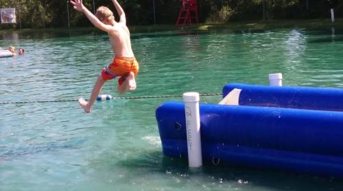 A boy in orange swim trunks jumps off a blue inflatable platform into a clear, greenish water.