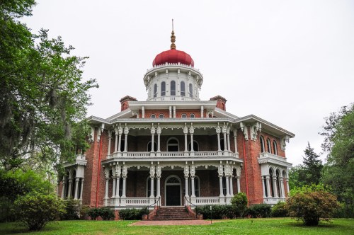 A grand, historic mansion with a red dome, intricate white trim, and lush greenery surrounding it.