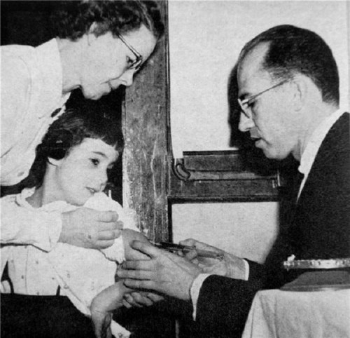 A child receives a vaccination from a doctor while a woman assists, all indoors.
