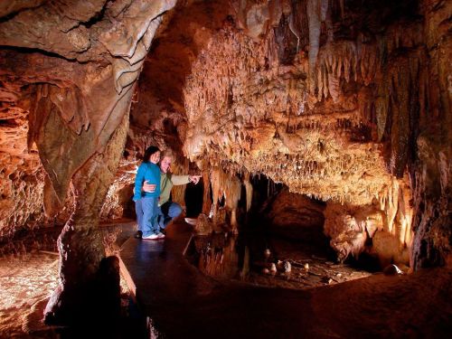 Two people explore a cave, surrounded by stunning stalactites and stalagmites, with water reflecting the cave's features.