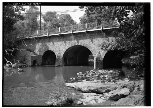 Black and white image of a stone bridge with arches over a calm river, surrounded by trees and rocks.