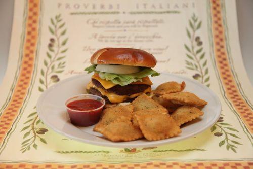 A cheeseburger with lettuce and onion on a plate, accompanied by ravioli and marinara sauce, set against a decorative background.