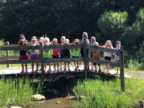 A group of children on a wooden bridge, each holding a camera, surrounded by greenery and a small pond.