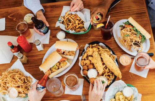 A table filled with various dishes, including sandwiches, fries, fried seafood, and drinks, surrounded by hands reaching for food.