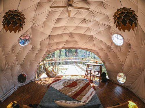 Cozy geodesic dome interior with wooden floor, large windows, a hammock, and modern decor overlooking nature.