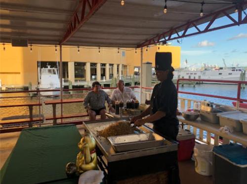 A chef prepares food on a deck overlooking a marina, with two diners seated nearby enjoying the view.
