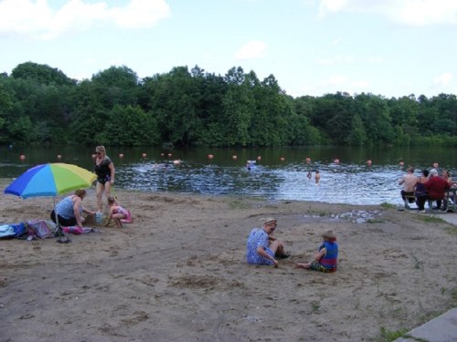 A sandy beach by a lake with people relaxing, swimming, and enjoying a sunny day under an umbrella.