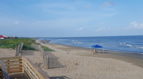 A sandy beach with gentle waves, a blue sky, and a few people near a blue tent in the distance.