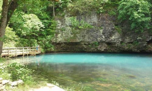 A serene blue pond surrounded by lush greenery and rocky cliffs, with a wooden bridge in the foreground.