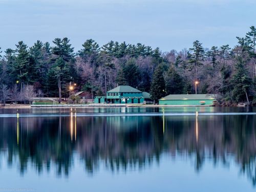 A serene lake scene with green buildings reflecting on calm water, surrounded by trees under a twilight sky.