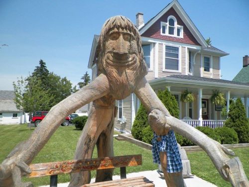 A large wooden sculpture of a smiling figure with outstretched arms, set in front of a house and green lawn.