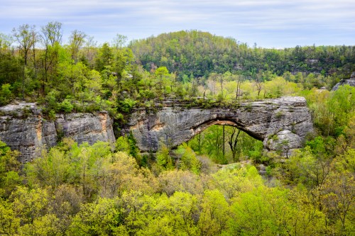 A natural stone arch surrounded by lush green trees and hills under a cloudy sky.