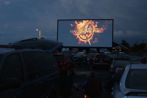 A drive-in movie screen displays "Catching Fire" as cars are parked in front under a twilight sky.
