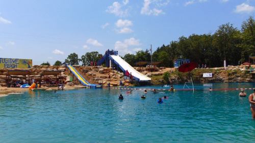 A sunny water park scene with a blue lake, water slides, and people swimming and relaxing around the shore.