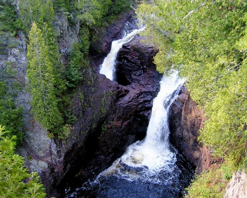 A scenic view of two waterfalls cascading over rocky cliffs, surrounded by lush green trees.