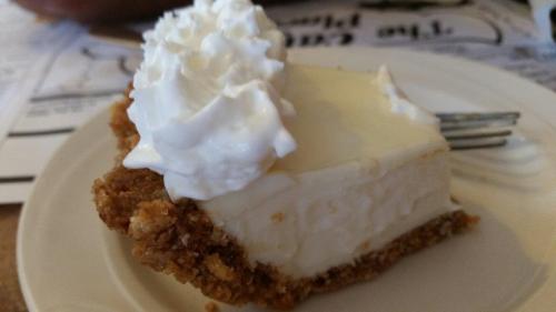 A slice of creamy dessert with a graham cracker crust, topped with a dollop of whipped cream on a white plate.