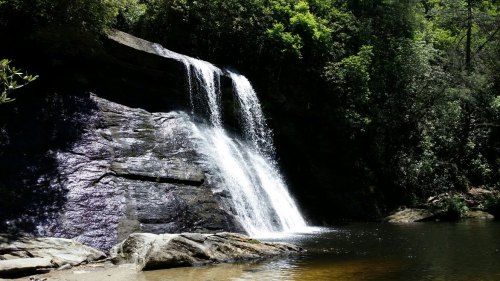 A serene waterfall cascading over rocks, surrounded by lush green trees and a calm pool of water below.