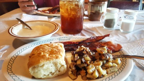 A plate with a biscuit, bacon, hash browns, and a bowl of soup, alongside a glass of iced tea.