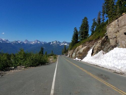 A scenic mountain road with snow patches, flanked by trees and rocky terrain under a clear blue sky.
