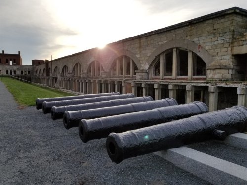 A row of historic cannons lined up in front of an old stone building, with the sun setting in the background.